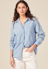 Camisa de mezclilla azul claro con botones en la parte delantera, bolsillo en el pecho y mangas remangadas. Usada con pantalones blancos, que tienen una textura suave y lisa.