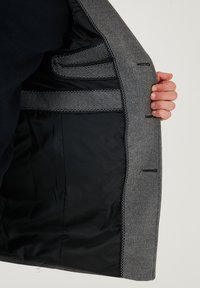 Interno di un blazer di lana grigia con una fodera nera liscia, dotato di diversi taschini. Cuciture visibili in bianco. Mano che tiene il blazer.
