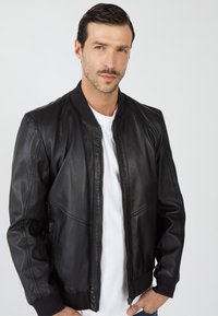 Zwarte leren bomberjack met een soepele textuur, geribde manchetten en kraag. Voorzien van een rits aan de voorkant en zijzakken. Draagt over een wit shirt.