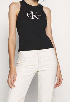 Kobieta ubrana w czarny bezrękawnik Calvin Klein Jeans i jasnobeżowe spodnie z wysokim stanem, stojąca z rękami swobodnie opuszczonymi wzdłuż ciała.