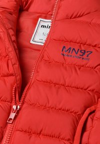 Röd puffjacka med horisontell quiltad textur, full dragkedja och en etikett med varumärket "MN97 NEVER STOP 97" på insidan.