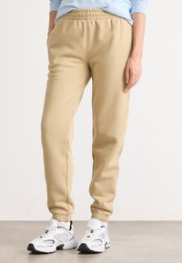 Beiga sweatpants med tapered fit, elastisk midja och sidofickor, i kombination med vita sportskor som visar nät- och gummidetaljer.