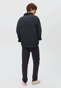 Čierna bavlnená bunda s vysokým golierom, výraznými švami a dlhými rukávmi, doplnená o tmavé nohavice a svetlé tenisky.