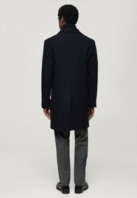 Long dark navy coat made of wool, featuring a structured collar and single vent at the back. Paired with gray trousers and black shoes.