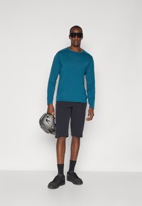 Langarmshirt in Teal, schwarze Shorts, schwarze Socken und Turnschuhe. Hält einen grauen Helm mit gemustertem Design und trägt große schwarze Sonnenbrillen.