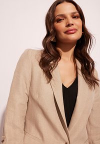Beżowa lniana marynarka o dopasowanym kroju, z klapami i zapięciem na jeden guzik, noszona na czarnym prążkowanym topie.