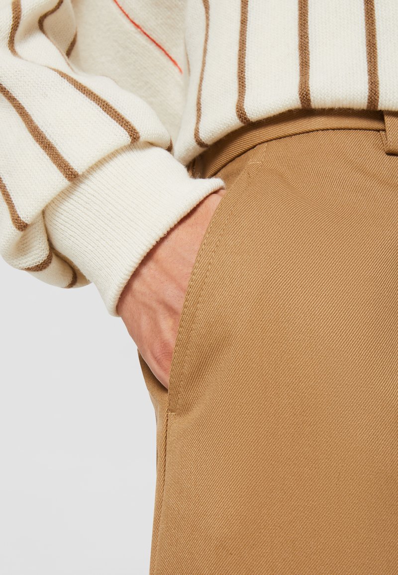 Pantalones de algodón beige con un bolsillo lateral, combinados con un suéter color crema con rayas marrones y un acento rojo. Primer plano de la mano en el bolsillo.
