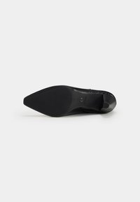 Chaussure à talon haut noire à bout pointu vue de la semelle avec une surface antidérapante texturée et un petit logo près de la voûte plantaire sur un fond uni.