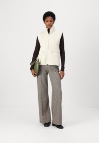 Vit fleeceväst med hög krage, dragkedja och två framfickor. Kombinerad med rutiga, vida byxor och en grön handväska.