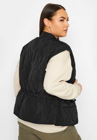 Svart vadderad väst med åtdragen midja, med rund krage och sidofickor. Buren över en ljus krämfärgad sweatshirt.