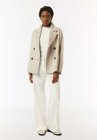 Light beige double-breasted coat with a notched collar, two large black buttons, paired with a white top and wide-leg cream trousers.