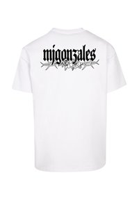 Balta kokvilnas t-kreklu ar melnu grafisku druku aizmugurē, kurā redzams vārds "migon zales", ko ieskauj ziedu un ērkšķu stieņu motīvi.