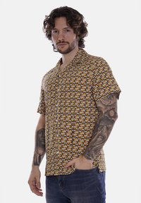 Camicia a maniche corte marrone con un motivo geometrico, caratterizzata da forme circolari. Realizzata in tessuto liscio, dotata di colletto e chiusura con bottoni.