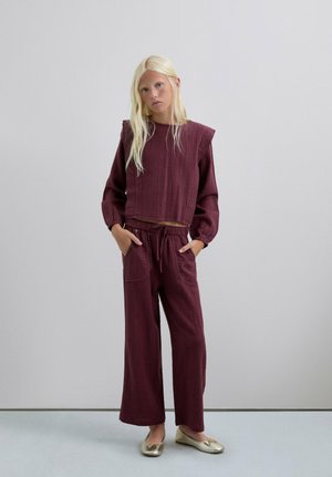 Conjunto burdeos de dos piezas con un top corto de tela texturizada y mangas abullonadas, combinado con pantalones de pierna ancha y zapatos planos dorados.