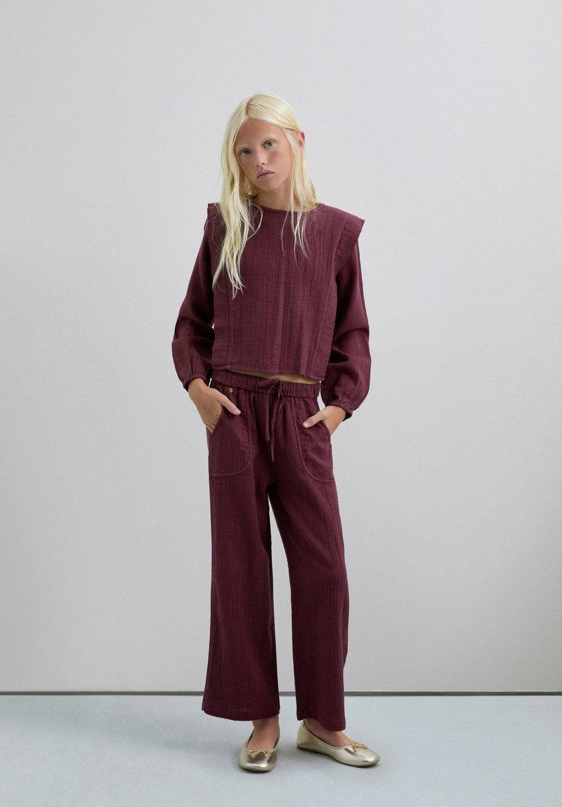 Conjunto burdeos de dos piezas con un top corto de tela texturizada y mangas abullonadas, combinado con pantalones de pierna ancha y zapatos planos dorados.