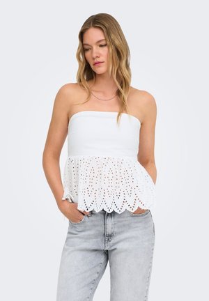 Femme aux longs cheveux blonds portant un top bustier blanc à œillets et un jean bleu clair, debout les mains dans les poches devant un fond uni.