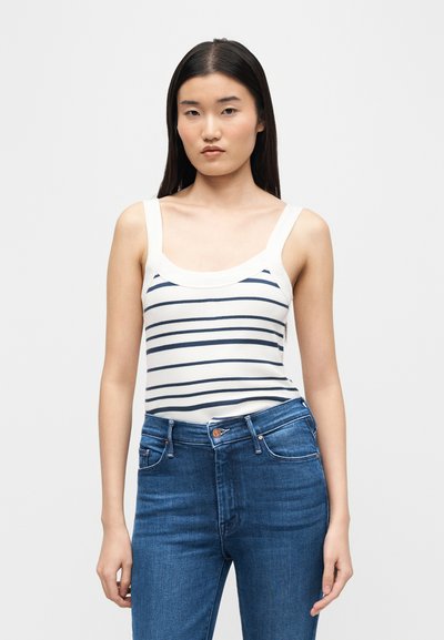 Γυναίκα με μακριά μαύρα μαλλιά φοράει λευκό τοπ με μαύρες οριζόντιες ρίγες και ψηλόμεσα μπλε τζιν, στέκεται μπροστά σε απλό φόντο.