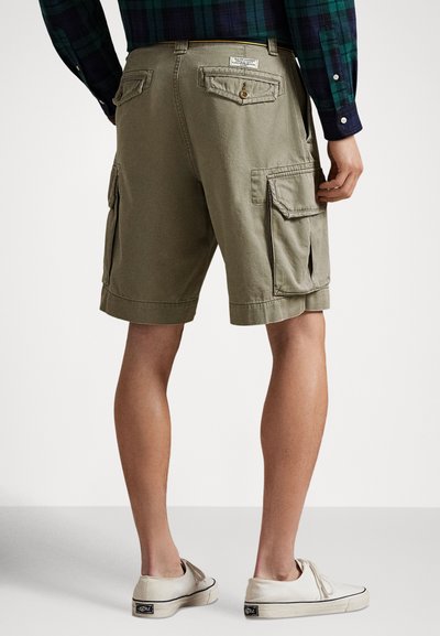 Kaki olivově zelené kraťasy s několika kapsami, včetně bočních a zadních, a ohrnutým lemem. Nošeny s světle barevnými plátěnými botami.