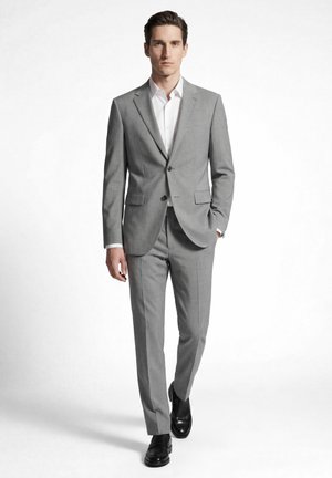 Man loopt, draagt een lichtgrijs tweeknops pak, wit overhemd en zwarte nette schoenen tegen een effen witte achtergrond.