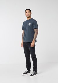 Čovjek s tetovažama na rukavu, obučen u tamnoplavu majicu s grafikonom orla, crne traperice i crne tenisice s bijelim potplatom, stoji na bijeloj pozadini.