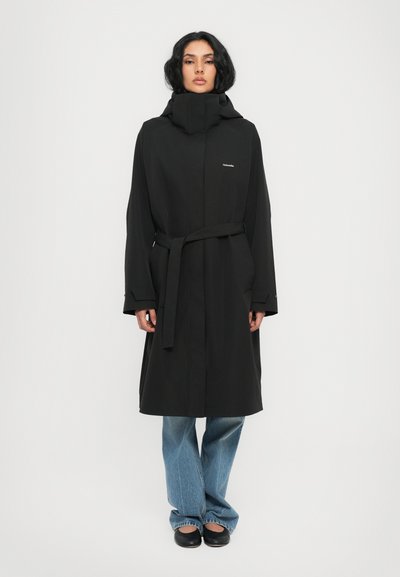 Sort lang frakk med høj krave, belagt talje og to sidelommer. Lavet af et glat materiale, parret med lyseblå flagrende jeans og sorte sko.