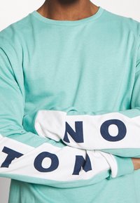 Světle tyrkysová košile s dlouhým rukávem a bílými rukávy, se tmavě modrým textem. Vyrobena z měkké tkaniny, hladká textura, uvolněný střih.