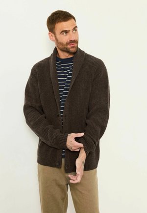 Mann mit kurzem Bart, der eine dunkelbraune Strickjacke über einem gestreiften Hemd und beigen Hosen trägt, und dabei den Ärmel der Strickjacke anpasst, während er seitlich schaut.
