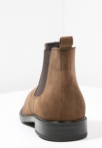 Botín de ante marrón con paneles laterales elásticos, tirador en la parte trasera y suela de goma. Presenta detalles de costura y un diseño de punta redonda.