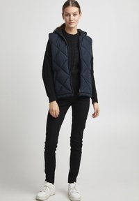 Navy padded vest with a hood, featuring diamond quilting. Worn over a black sweater, paired with black slim-fit pants and white sneakers.