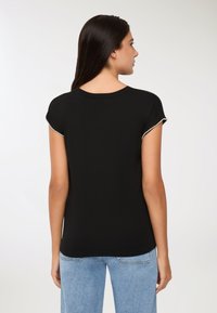 Kobieta z długimi ciemnymi włosami, ubrana w czarny krótki top z koralikowymi wykończeniami na rękawach i jasnoniebieskie dżinsy, widok z tyłu.