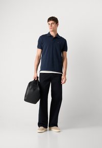 Marineblå polo med korte ærmer, slim fit og et lille logo. Sort taske med et robust håndtag. Mørke denimbukser og beige sko.