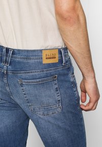 Denimjeans i medelblått med en texturerad yta. Har en läderlapp med texten "BLEND" och en distinkt design på bakfickan.