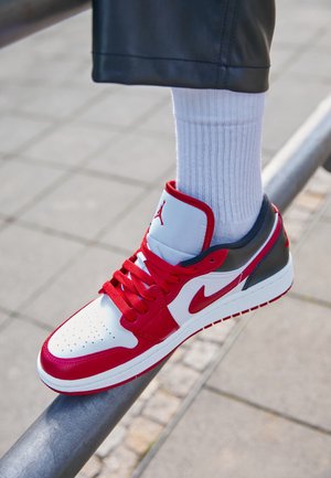 Persoon die rode, witte en zwarte Nike Air Jordan sneakers en witte sokken draagt, balancerend met één voet op een metalen reling buiten op een geplaveid oppervlak.