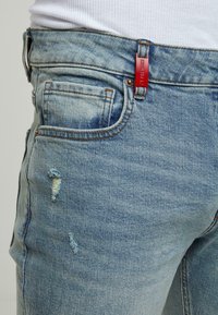 Světle modré džíny z denimu s poškozenými detaily, viditelným šitím a červenou nášivkou na pasu. Klasický design se pěti kapsami.
