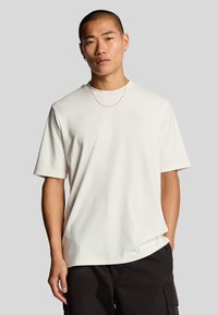 Kortærmet, krämfarvet t-shirt med en glat tekstur og prægede detaljer på brystet, sat sammen med mørke cargo-bukser.