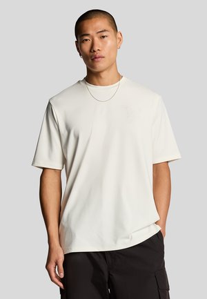 Kortärmad, krämfärgad t-shirt med en slät textur och präglad design på bröstet, tillsammans med mörka cargobyxor.