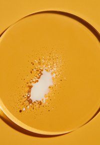 Round, clear glass plate on a textured, bright yellow background. The plate contains a swirl of brown liquid and white foam accent.