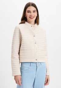 Beige gewatteerde jas met een ronde kraag, zilveren knopen en twee voorzakken. Draagt met lichtblauwe spijkerbroek.