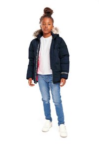 Giacca puffer blu navy con dettagli in pelliccia sintetica sul cappuccio, chiusura con zip e accenti del logo sulle maniche. Abbinata a una maglietta bianca e jeans blu.