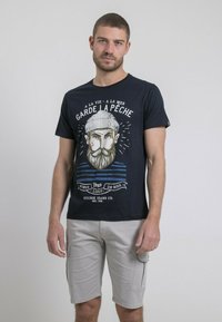 T-shirt en coton bleu marine avec un graphisme d'un homme barbu portant un chapeau, arborant des accents rayés blancs et bleus et le texte "GARDE LA PÊCHE." Associé à un short cargo gris.