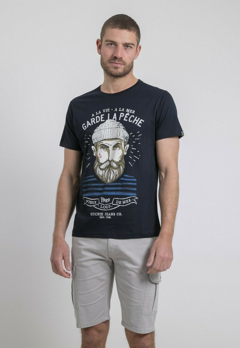T-shirt en coton bleu marine avec un graphisme d'un homme barbu portant un chapeau, arborant des accents rayés blancs et bleus et le texte "GARDE LA PÊCHE." Associé à un short cargo gris.