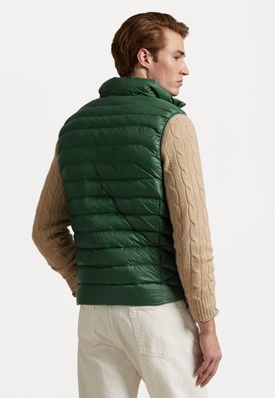 Gilet matelassé vert avec texture matelassée horizontale, col haut et fermeture éclair, associé à un pull en tricot beige et un pantalon de couleur claire.