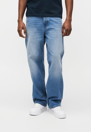 Uomo che indossa jeans denim azzurri larghi, sneakers bianche e una maglietta nera, in piedi su sfondo bianco.