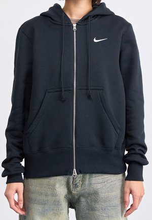 Osoba ubrana w czarną bluzę z zamkiem i kapturem marki Nike z przednimi kieszeniami i troczkami, w połączeniu z wyblakłymi niebieskimi dżinsami.
