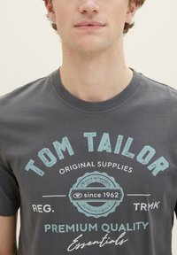 Grå bomulds t-shirt med rund hals, korte ærmer og trykt tekst i lys blå og hvid, inklusive "TOM TAILOR" og "siden 1962."
