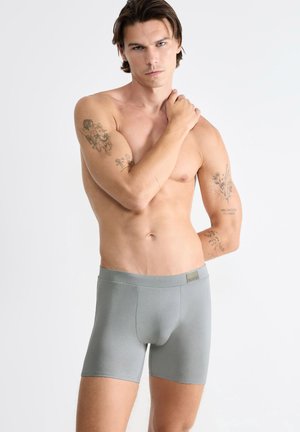 Skjorteløs mand med tatoveringer på begge arme iført grå boksershorts, står mod en ensfarvet hvid baggrund og rører ved sin skulder.