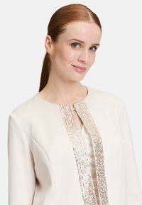 Jasno beżowa kurtka z wyszywaną cekinami przednią częścią oraz eleganckim, minimalistycznym wzornictwem. Materiał wydaje się gładki z wyraźnie zarysowanymi ramionami.