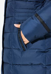 Marineblå puffjacka med quiltad textur, med en dragkedjeficka på vänster sida och svart accentrand på ärmen.