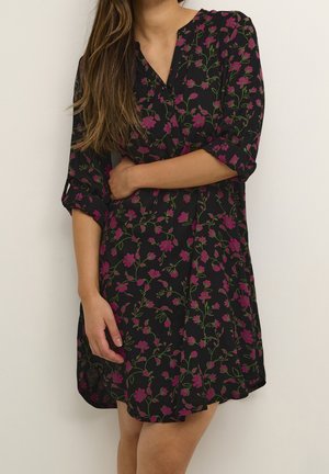 Frau trägt ein schwarzes Blumenkleid mit pinken Blumen und grünen Stielen, die Ärmel hochgekrempelt, posiert mit einem Arm über dem Körper.