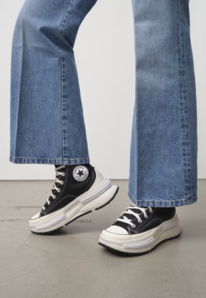 Henkilö, jolla on siniset leveälahkeiset farkut ja mustavalkoiset korolliset Converse All Star -tossut harmaalla lattialla.